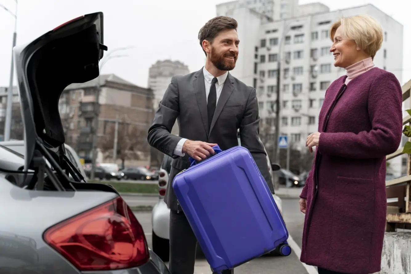A professional chauffeur in a suit loads a purple suitcase into the trunk of a black car while smiling and interacting with a female client, representing Kingston Ontario limo service.