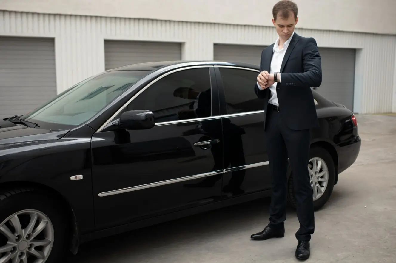 Professional chauffeur checking time beside black luxury sedan for London Ontario limo service pickup outside building